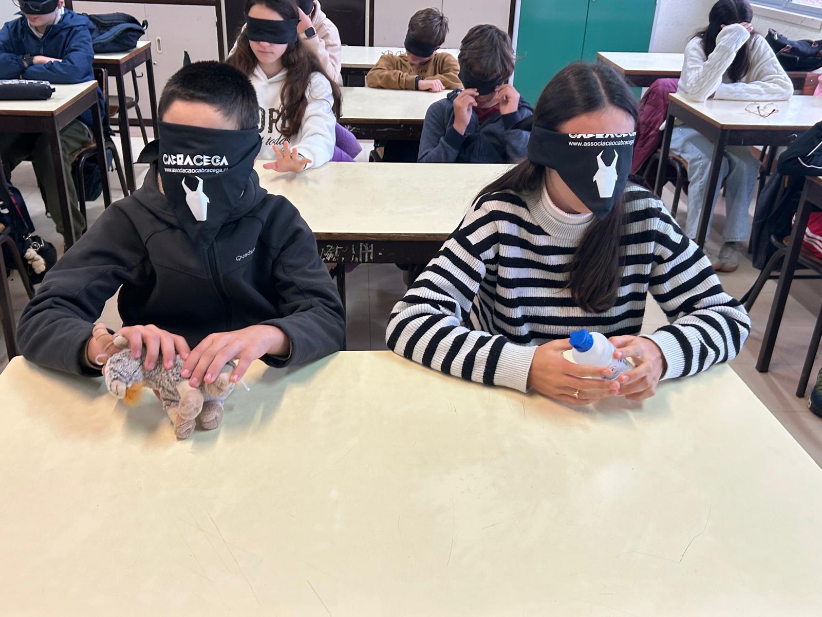 Uma sala de aula com vários adolescentes sentados em carteiras, todos com vendas pretas nos olhos.  Em primeiro plano, há dois alunos na mesa da frente: À esquerda, um aluno com uma camisola de fato de treino escura usa uma venda com o texto “CABRA CEGA” e está a tocar, com as duas mãos, num pequeno animal de peluche cinzento (uma cabra). À direita, uma aluna com uma camisola de malha às riscas pretas e brancas também usa uma venda “CABRA CEGA” e segura com as duas mãos uma pequena garrafa de álcool etílico.  Ao fundo, vêem-se outros alunos igualmente vendados, a participar na mesma atividade, alguns parecem estar a ajustar a venda ou com as mãos junto ao rosto. À direita há um armário e a luz parece vir das janelas. 